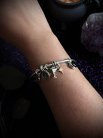 A silver charm bracelet with a martini glass, star, and crescent moon design, worn on a person's wrist against a dark, mystical background with purple glitter and crystals.