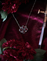 Silver necklace with a red gemstone pendant on a burgundy fabric background with flowers and a sword.