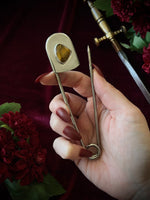 Hand holding a giant steel safety pin with a tiger's eye gemstone against a dark background with flowers and a sword.