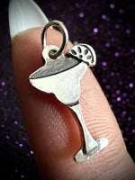 A silver-toned cocktail glass charm with a lemon wheel detail, shown resting on a person's finger against a dark purple glittery background.