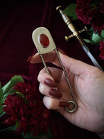 Hand holding a giant steel safety pin with a red gemstone against a dark background with flowers and a sword.