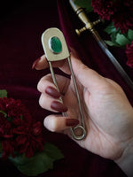 Giant stainless steel safety pin with a green gemstone on a dark burgundy fabric background with flowers and a sword.