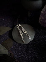 A pair of silver-toned dangling earrings with crescent moon, star, and bird motifs, displayed on a dark stone slab against a dark, sparkly background with decorative leaves and crystals.