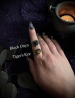 A hand with dark manicured nails displays two heart-shaped rings on the fingers. One ring features a black onyx stone, and the other features a tiger's eye stone, set in silver-toned metal against a dark, sparkly purple background with decorative leaves and a small candle.