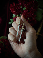 Hand holding a pair of sword-shaped earrings with a dark floral background.
