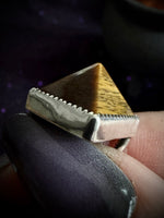 A close-up of a ring being held between fingers. The ring features a triangular pyramid-shaped face with a polished golden-brown top and a textured silver-toned band with a serrated edge.