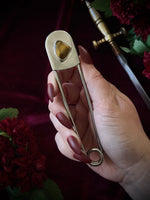 Hand holding a giant steel safety pin with a tiger's eye gemstone against a dark background with flowers and a sword.