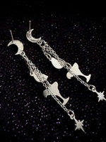 A pair of silver-toned dangle earrings featuring crescent moon, wine glass, and star motifs connected by delicate chains, displayed on a dark glittery background.