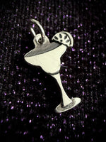 A silver-toned metal charm shaped like a margarita glass with a lime wheel garnish, shown on a dark glittery purple background.