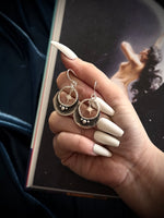 Hand holding a pair of silver hoop earrings with a star design, against a dark background.