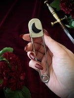 Hand holding a giant steel safety pin with a black crescent moon gemstone against a dark background with flowers and a sword.