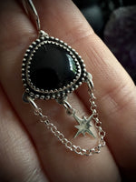 A close-up of a hand holding a silver-toned earring with a large black teardrop-shaped stone and a dangling star charm on a delicate chain.