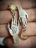A pair of dangling hand-shaped earrings held against a person's fingers. One earring is silver-toned with a sunburst at the top and a larger openwork hand dangle, while the other is gold-toned with a crescent moon dangle.
