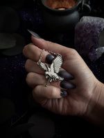 A hand with dark purple nail polish holds a silver bird pendant on a chain against a dark, mystical background with a cauldron and amethyst crystal.