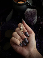 A hand with dark gradient stiletto nails wearing a small silver fairy ring charm, set against a dark, mystical background with an amethyst cluster and a cauldron.