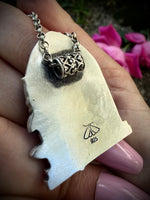 Hand holding the backside of a silver necklace with intricate design and pink flowers in the background