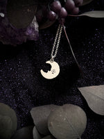 A silver crescent moon pendant necklace with a custom engraved character, displayed on a dark glittery background with dried leaves and purple crystals.