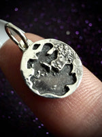 A close-up of a silver pendant with an organic, textured shape held on a person's finger against a dark purple glittery background.