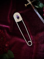 Giant stainless steel safety pin with an amethyst gemstone on a dark burgundy fabric background with flowers and a sword.