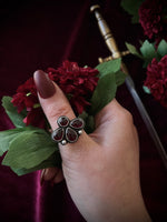 Hand wearing a decorative gemstone ring with red stones against a dark background with flowers and a sword.