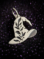 A silver witch hat pendant with leaf cutouts, displayed on a sparkling black background with purple highlights.