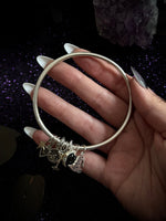 A silver-toned bangle bracelet with celestial charms, held in a hand with long white manicured nails against a dark glittery background with a purple crystal.