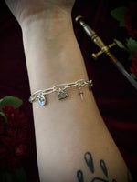 Silver chain bracelet with mixed metal charms on a wrist against a dark background with flowers and a sword.