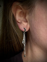 A close-up of a person's ear wearing a silver crescent moon hoop earring with a dangling chain, skull, and star charm.