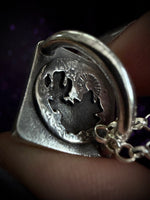 A close-up of a silver-toned pendant necklace being worn on a finger. The pendant features a textured, organic shape with a circular inset showing a stylized sun and moon design, with a silver chain visible on the right.