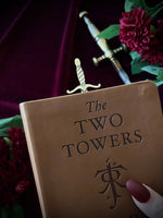 Leather book titled 'The Two Towers' with a sword bookmark sticking out of the top, against flowers and a sword in the background.