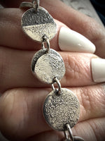 Silver circular pendant on a chain held between fingers with white nail polish.
