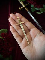A hand holding a sword-shaped bookmark on a dark background surrounded by flowers and a sword.