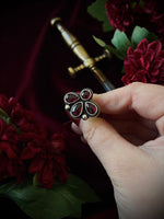 Hand holding a decorative gemstone ring with flowers and a sword in the background.