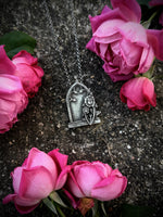 Silver gothic gravestone pendant on a silver chain against a dark, stone surface surrounded by pink roses.