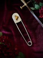 Giant stainless steel safety pin with a tiger's eye gemstone on a dark burgundy fabric background with flowers and a sword.