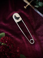 Giant stainless steel safety pin with a black crescent moon gemstone on a dark burgundy fabric background with flowers and a sword.