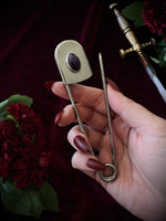 Hand holding a giant steel safety pin with an amethyst gemstone against a dark background with flowers and a sword.