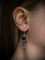 A close-up of a person's ear wearing a silver-toned dangle earring with a black oval stone and a dangling star charm.