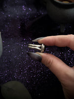 A hand with long dark glittery nails holds a silver-toned ring with a textured, patterned band over a dark purple glittery background.