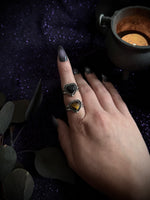 A hand with long dark gray manicured nails wearing two stacked silver rings, one with a black heart-shaped stone and the other with a yellow heart-shaped stone, posed on a dark purple glittery surface next to a candle.