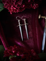 Silver and brass sword earrings on a burgundy fabric background surrounded by a sword and flowers.