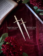 Two decorative silver sword bookmarks on a dark fabric background surrounded by flowers, a sword, and an open book.