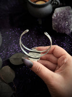 A hand with dark glittery nail polish holds a silver-toned triple-band cuff bracelet against a dark, mystical background with purple glitter, a cauldron, and an amethyst cluster.