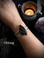 A silver-toned double-band cuff bracelet with an oblong black onyx stone, shown worn on a person's wrist. The background is dark and mystical, featuring a lit candle in a small pot, a purple crystal cluster, and the text 'Oblong' overlaid on the bottom left.