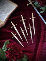 Set of decorative silver sword bookmarks on a burgundy fabric background, surrounded by flowers, a sword, and an open book.