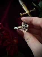 Silver ring with a decorative design held by a hand against a dark background with red flowers and a sword.