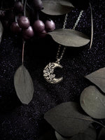 A silver crescent moon pendant necklace with delicate leaf filigree, displayed on a dark textured background with dark green leaves and purple berries.