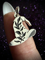 A sterling silver witch hat pendant with leaf cutouts, held on a finger against a dark purple glittery background.