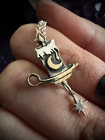 A close-up of a hand holding a silver and gold pendant shaped like a melting candle with a dangling star charm.