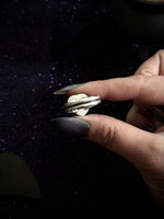A close-up of a hand with long dark glittery nails holding a silver ring with a small white heart-shaped stone against a dark, sparkly background.